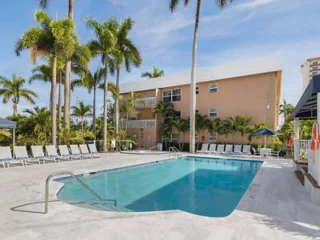 Outdoor swimming pool with lounge chairs and umbrellas, surrounded by palm trees and a multi-story building in the background on a sunny day.