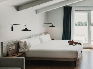 A neatly made double bed in a minimalist bedroom with wall-mounted lamps, a window with dark curtains, and a small bag on the bed.