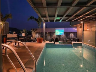 Rooftop pool area at night with illuminated water, metal handrails, lounge chairs, potted plants, and a movie projected on a screen in the background.