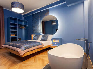 A modern blue bedroom with a round mirror, wooden bed frame, white bedding, a built-in shelving unit, and a freestanding white bathtub on a wooden floor.