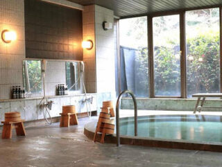 An indoor onsen with round hot tub, wooden stools, hand showers, and large windows offering a view of greenery outside.