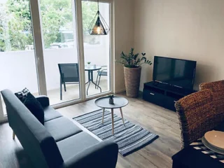 A modern living room features a gray sofa, a small coffee table, a flat-screen TV, and large windows opening to a balcony with two chairs and a table.