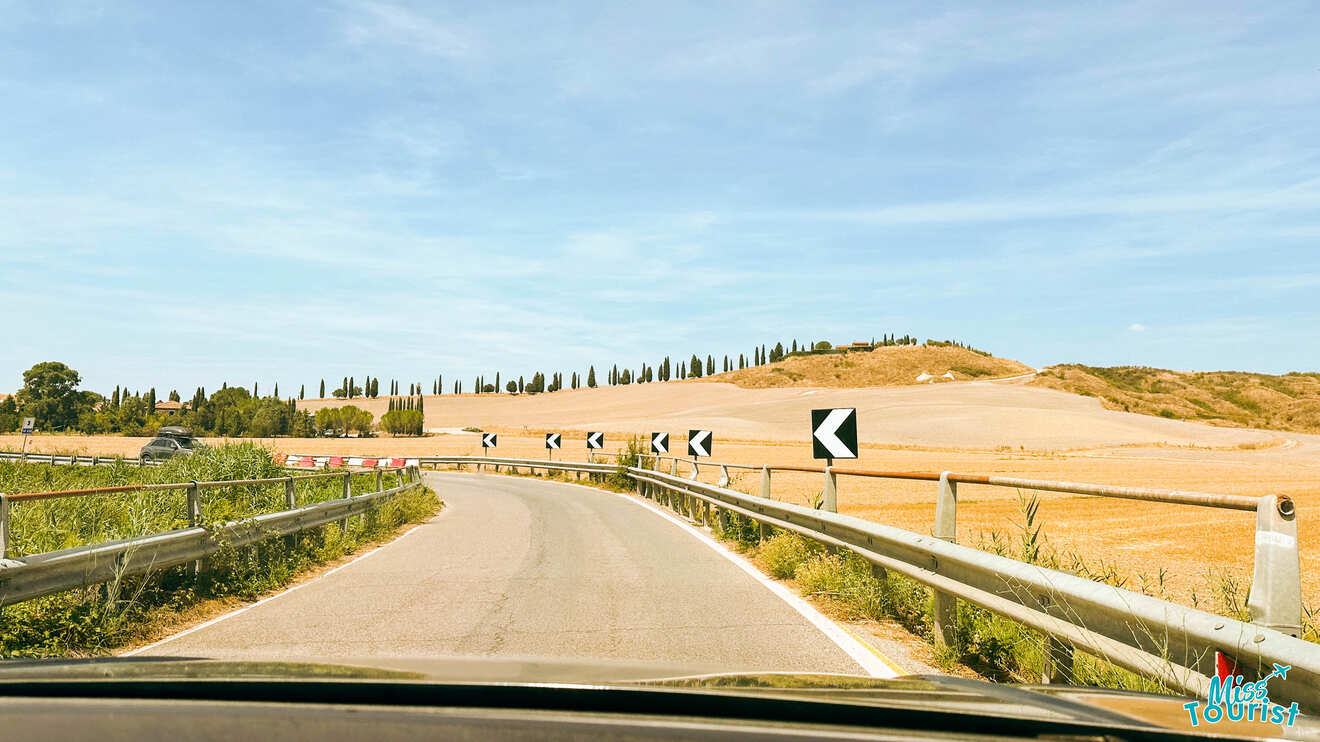 A road curves through a rural landscape with road signs indicating the direction of turns. Guardrails line the sides, and fields with sparse vegetation are visible under a clear sky.