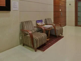 Two striped armchairs with wooden arms are placed beside a small round table holding magazines in a hallway. The floor is tiled and the walls are paneled.