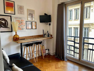 Small modern apartment features a high table with stools, wall-mounted TV, framed artwork, a potted plant, a couch, and large windows with gray curtains. The floor is wooden parquet.