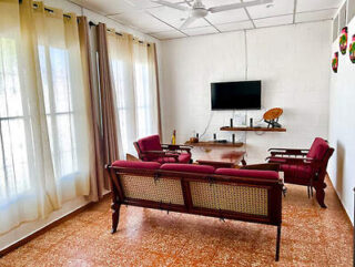 A living room with large windows, curtains, a wall-mounted TV, a shelf, and four red cushioned wooden chairs around a wooden coffee table on a tiled floor.