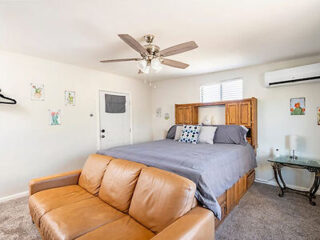 A bedroom with a ceiling fan features a neatly made bed with grey bedding, a wooden headboard, a brown leather sofa at the foot of the bed, and a small glass side table. Artworks adorn the white walls.