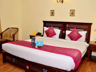 A neatly made bed with white sheets, pink accents, and two pink pillows. A small table on the left holds a menu and items in a room with warm lighting and simple decor.