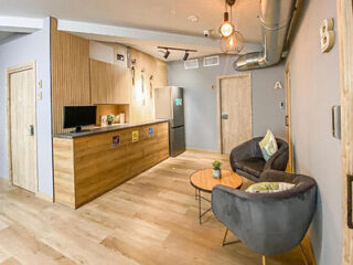 A modern kitchen/lounge area with wooden cabinetry, a stainless steel fridge, two armchairs, and a small round coffee table. The space has industrial design elements and warm lighting.