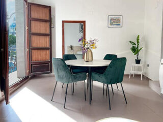 A dining area with a round white table and four green velvet chairs. An open wooden door leads to a balcony, and a plant and framed artwork decorate the white walls.