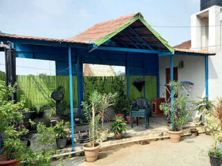 A small outdoor seating area with plants, a blue awning, and simple patio furniture at a guesthouse.