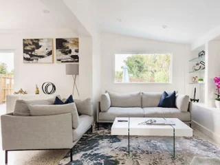 A modern living room with two gray sofas, a white coffee table, abstract wall art, a patterned rug, and a large window allowing natural light. Accessories include a floor lamp and decorative items.