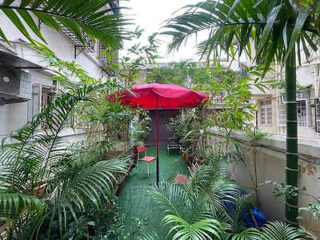 A narrow outdoor space with abundant greenery, featuring a red umbrella, two chairs, and a small table on a green artificial lawn.