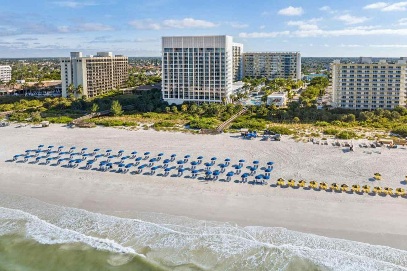 Aerial view of a sandy beach with rows of blue and yellow beach umbrellas, in front of several large hotels and buildings, with greenery in the background.