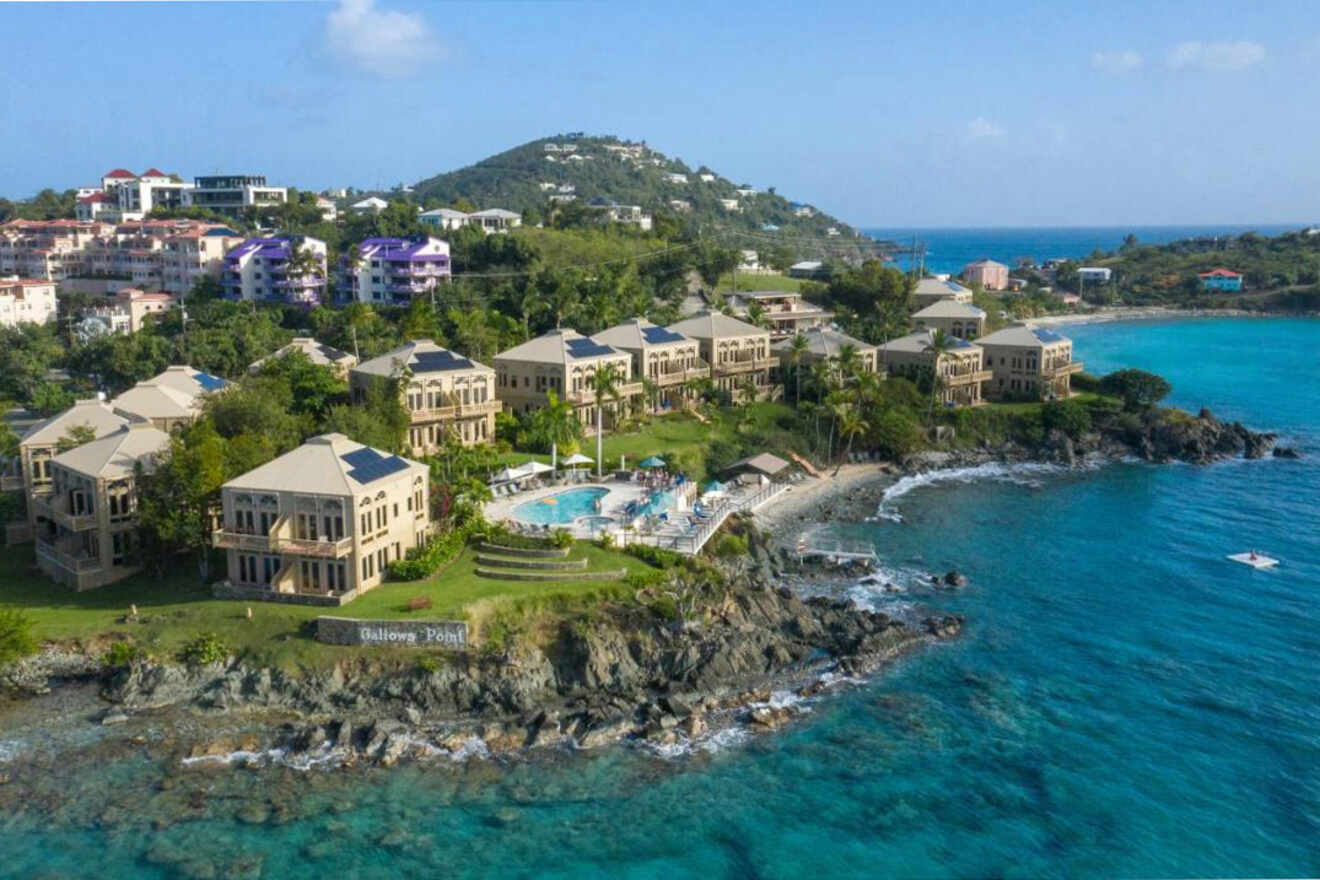 Aerial view of a coastal resort with multiple buildings on a rocky shoreline, a central swimming pool, and surrounding greenery, set against a backdrop of blue ocean and distant hills.