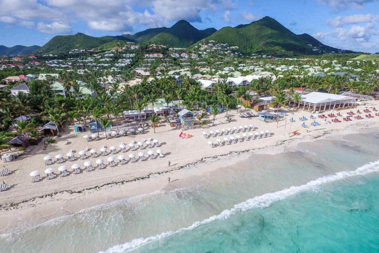 A picturesque beach lined with white umbrellas and lounge chairs, surrounded by palm trees and small houses, with green hills in the background.