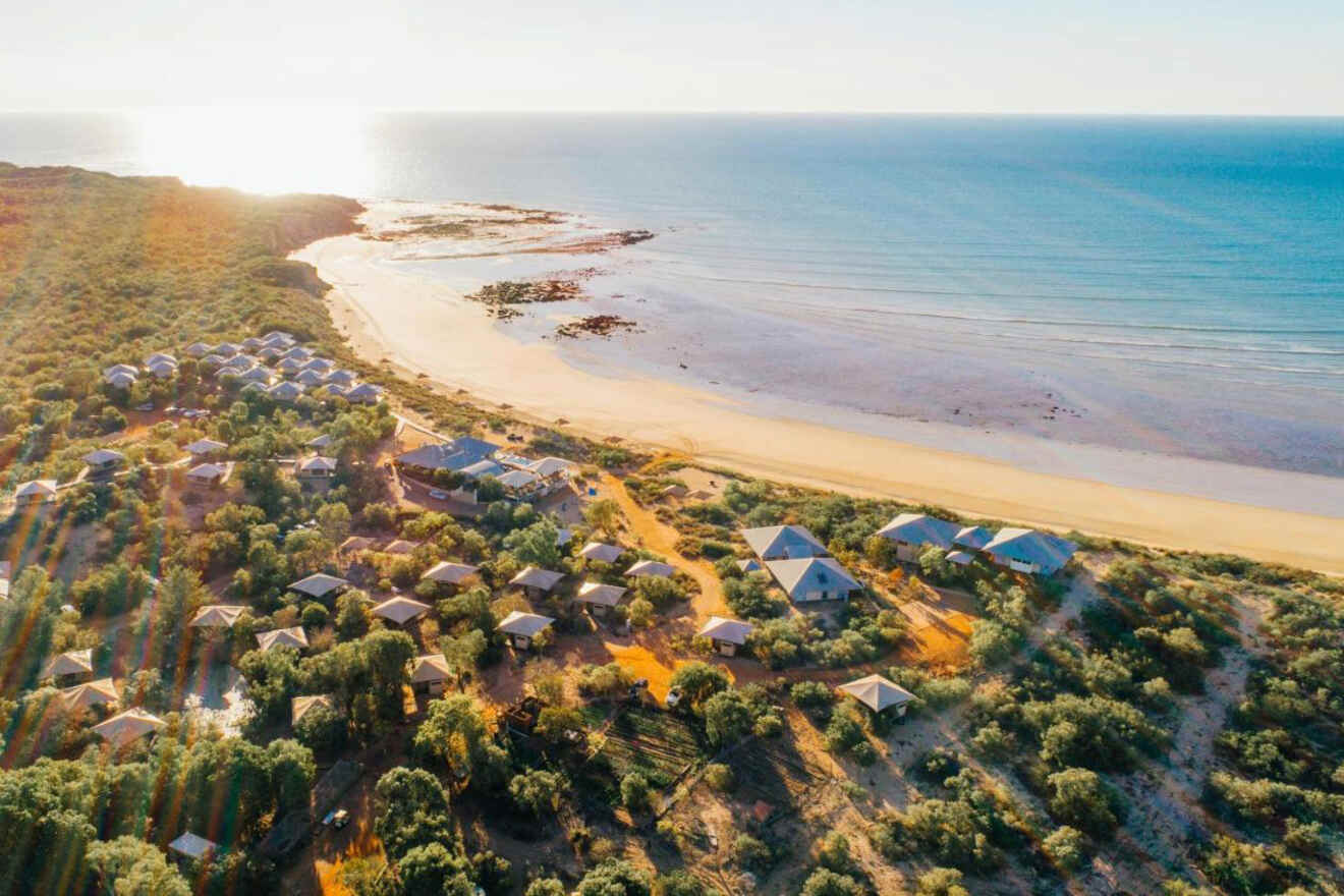 Aerial view of a coastal area featuring a beach, ocean, and a resort with various buildings surrounded by greenery, under a clear sky.