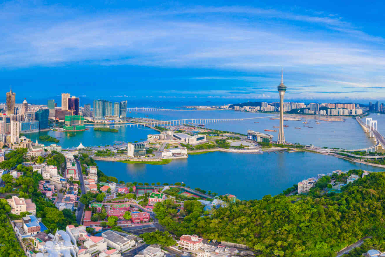 Panoramic view of the Macau skyline, featuring the Macau Tower, bridges, lush green hills, and the cityscape surrounded by water under a clear blue sky.