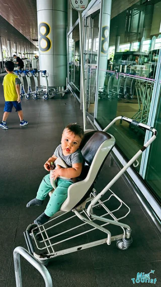 A child is sitting in a stroller at an airport terminal. A boy in yellow and blue clothing is walking in the background near a row of luggage trolleys.