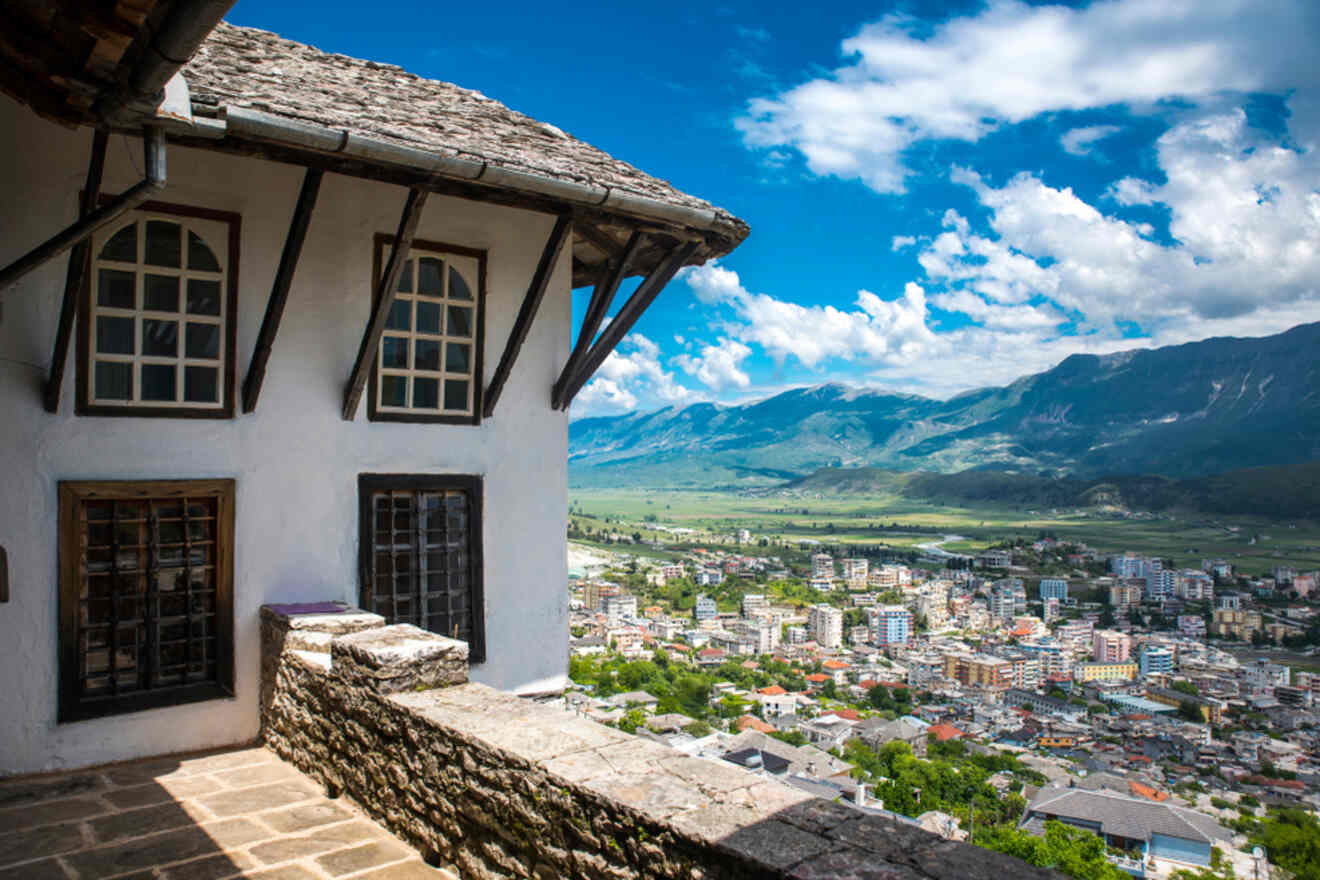 A balcony view in Gjirokastër, Albania, showing traditional architecture with wooden windows, overlooking the valley and surrounding mountains under a clear blue sky.