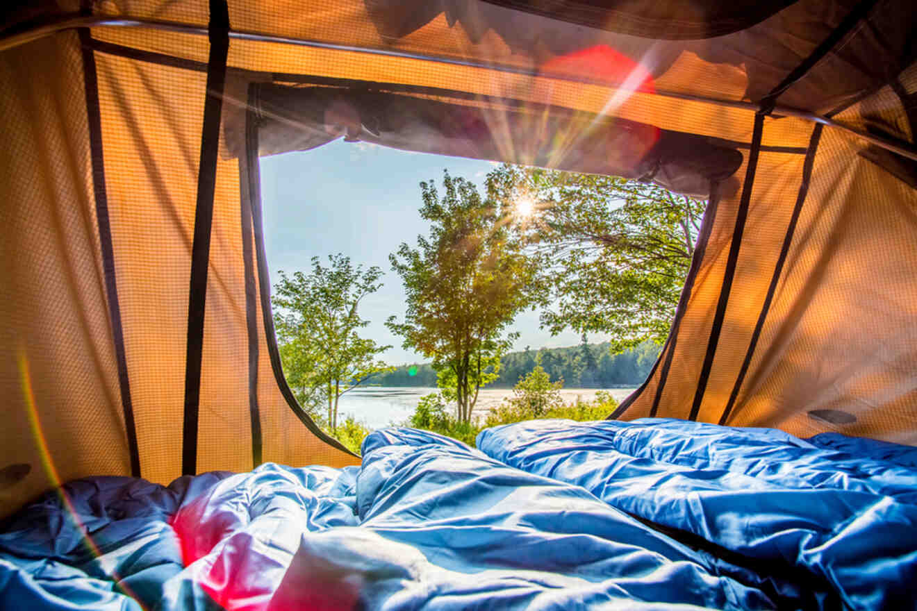 View from inside a tent showing the entrance unzipped, revealing a sunny outdoor scene with trees, a lake, and blue sleeping bags in the foreground.