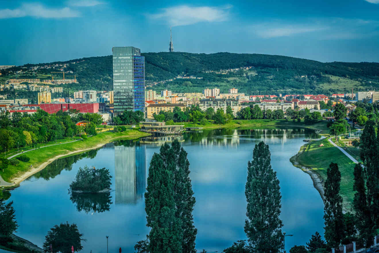 Cityscape with a lake surrounded by greenery, reflecting nearby buildings, a tall glass skyscraper, residential areas, and a hill with a tower in the background.