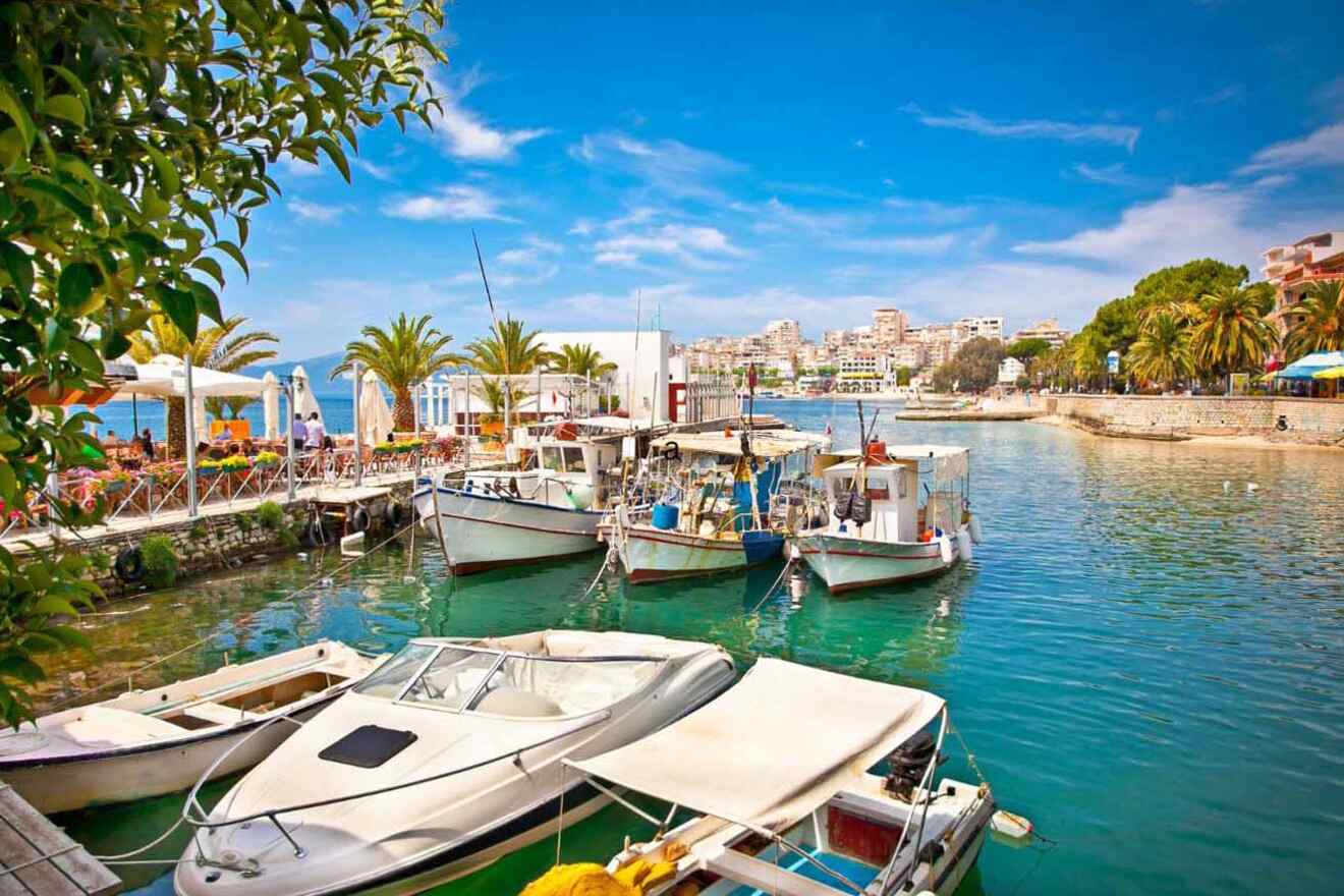 Small boats are docked in a vibrant harbor under a clear blue sky, with a backdrop of palm trees, buildings, and a few people sitting at outdoor cafes by the water's edge.