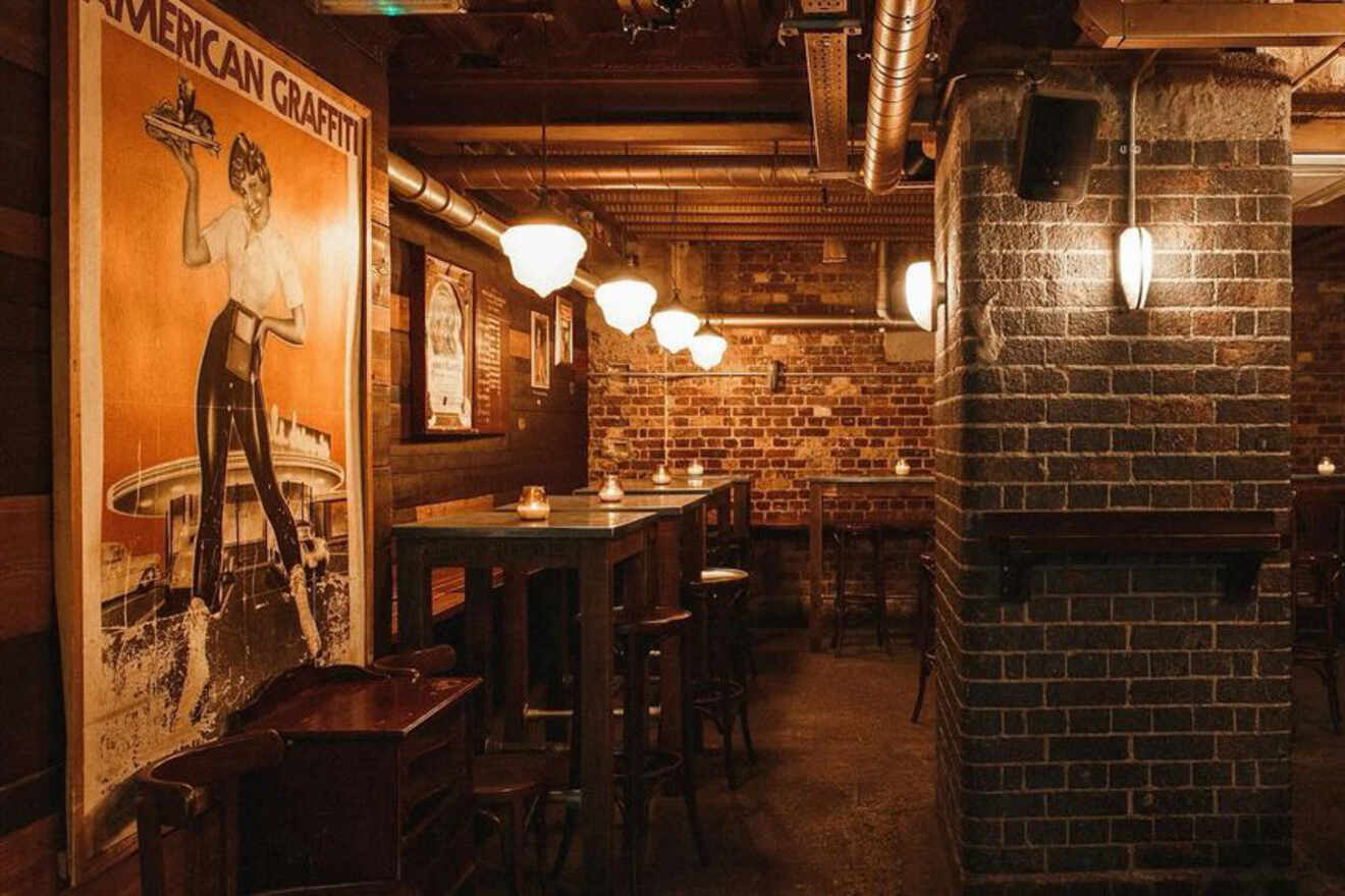 A cozy bar area with exposed brick walls and vintage décor, featuring a tall “American Graffiti” poster and dim lighting.