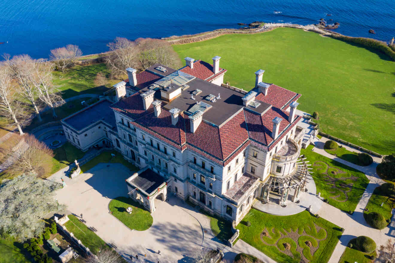 Aerial view of a large, historic mansion with a red roof, surrounded by manicured lawns and located near a body of water.