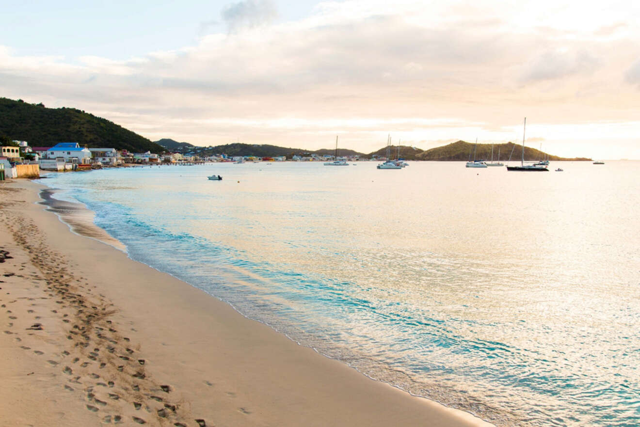 A peaceful beach at sunset, with calm waves lapping against the shore and boats anchored in the distance, set against a backdrop of hills and coastal homes.