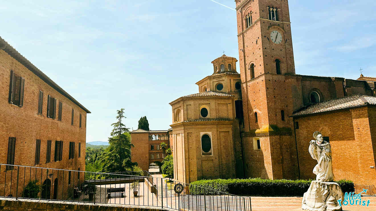 Sunny view of a historic Italian town square, featuring a large brick church with a bell tower and a statue of a religious figure in the foreground.