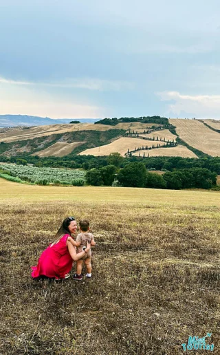 The writer of the post sits in a golden Tuscan field with her child, both smiling and enjoying a serene landscape dotted with cypress trees.