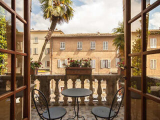 A quaint balcony with a small table and two chairs overlooks a courtyard surrounded by beige brick buildings and palm trees under a partly cloudy sky.