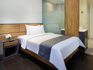 A neatly made hotel bed with white linens and a grey throw, next to a wooden nightstand with a flower arrangement. A frosted glass partition separates the bed area from the bathroom.