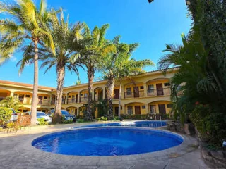 A circular swimming pool in the courtyard of a two-story building, surrounded by palm trees and greenery. Patio chairs and a table are on the left side of the pool. The sky is clear and blue.