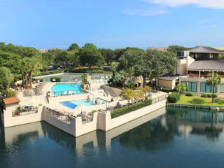 Aerial view of a resort complex featuring an outdoor pool and lounge area surrounded by greenery and overlooking a reflective water body.