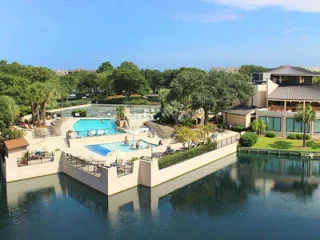 Aerial view of a resort complex featuring an outdoor pool and lounge area surrounded by greenery and overlooking a reflective water body.