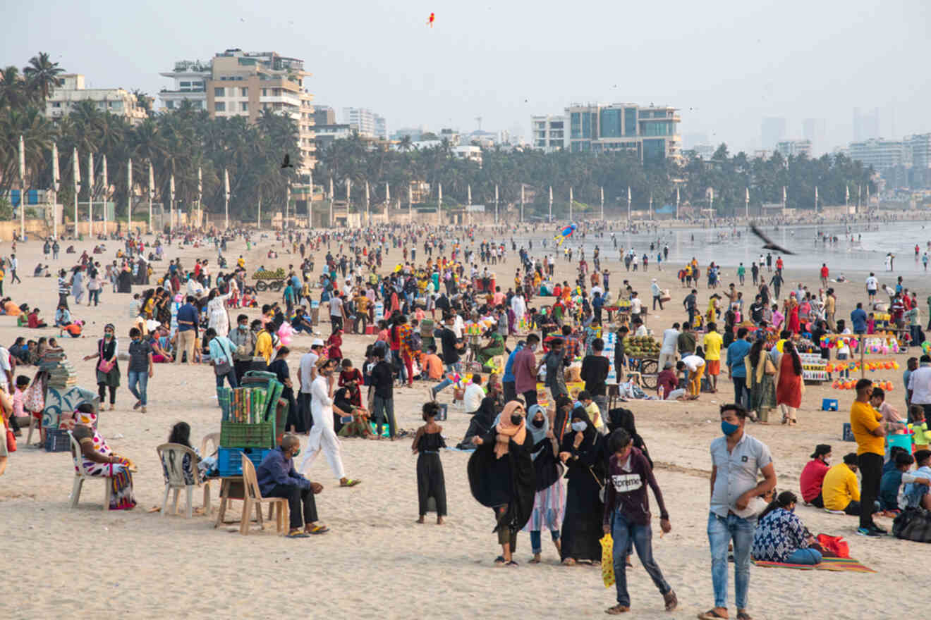 A crowded beach with people walking, sitting, and engaging in various activities. Many are seen wearing masks, with buildings and palm trees visible in the background.