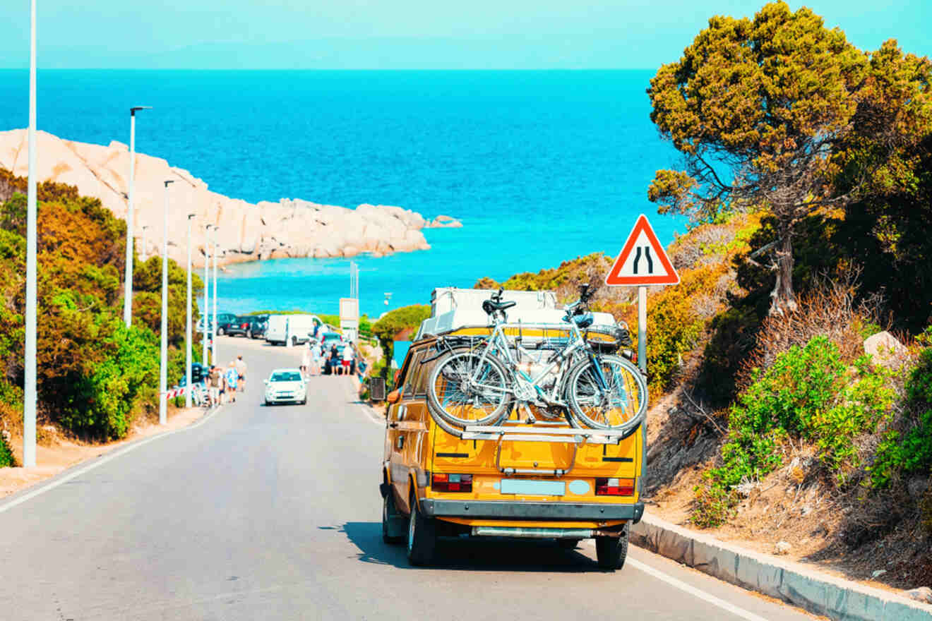 A yellow van with bicycles mounted on the back drives down a coastal road toward the sea, flanked by green bushes and rocks.