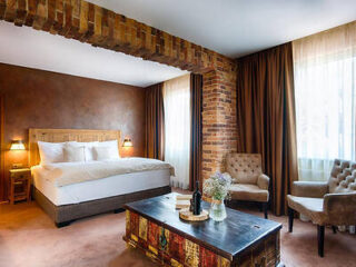 A warmly lit hotel room featuring a large bed, two upholstered chairs, a rustic coffee table with wine and flowers, and exposed brick accents.