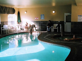 An indoor pool area with a person swimming in a hot tub, people seated on chairs by the pool, and a cascade of water flowing from a pipe into the main pool.