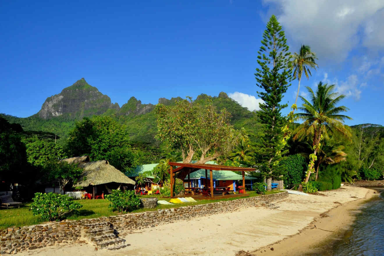 A sandy beach with a wooden gazebo and thatched hut, surrounded by lush vegetation, with mountains in the background under a clear blue sky.