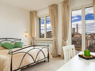 A bedroom with a metal-framed bed, two green pillows, a beige bedspread, a desk with a green candle, and two large windows with beige curtains offering a view of a cityscape.