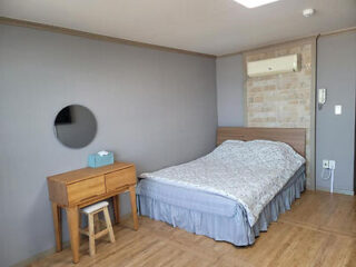 A minimalist bedroom with a bed, nightstand, circular mirror, wooden desk, and stool. The bed has a gray skirt and light-colored bedding. An air conditioning unit is mounted on the wall.