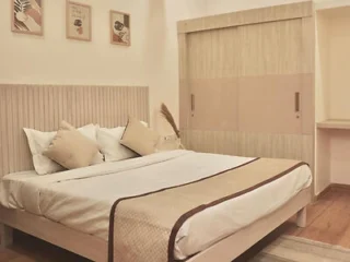 A neatly made bed with beige bedding and several pillows in a simple, modern bedroom featuring a wooden headboard, framed wall art, and a large built-in wardrobe.