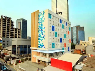A modern mid-rise building with a colorful blue and white geometric facade, located in an urban area with other tall buildings. A construction site is visible to the left.