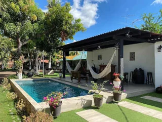 A backyard with a rectangular pool, hammocks under a covered patio, potted plants, and lush greenery. The sky is clear and blue.