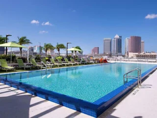 Rooftop pool with lounge chairs and umbrellas, overlooking a cityscape with tall buildings and clear blue skies.