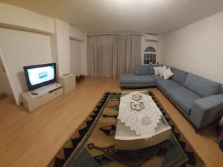 Living room with a blue sectional sofa, a TV on a stand, a patterned rug, and a coffee table with a lace tablecloth. White curtains cover the window.