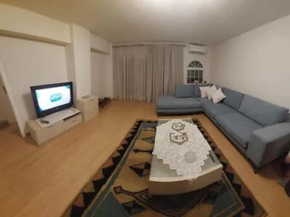 Living room with a blue sectional sofa, a TV on a stand, a patterned rug, and a coffee table with a lace tablecloth. White curtains cover the window.